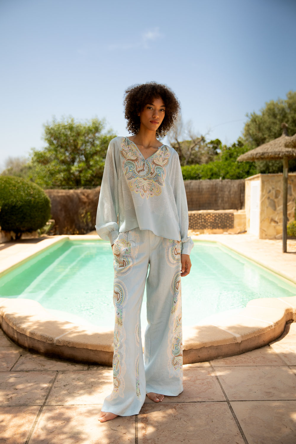 Imagebild: Model steht vor Pool und trägt EMBELLISHED LACE Seidenbluse und Leinenhose.