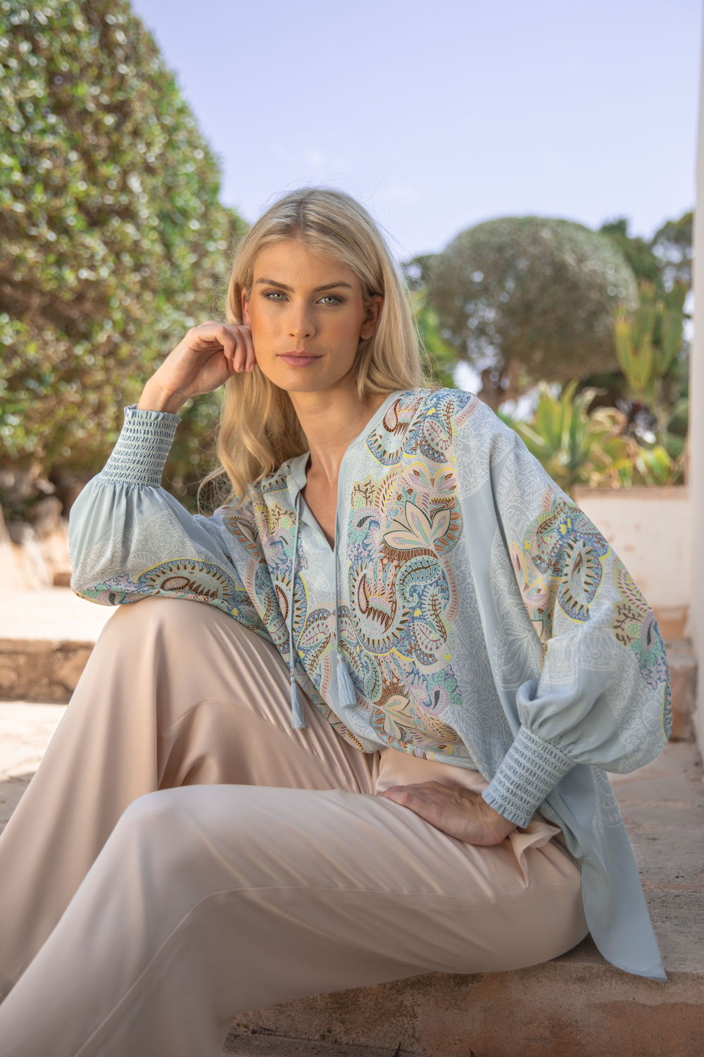 Imagebild: Model sitzt vor Finka in Mallorca und trägt EMBELLISHED LACE Tunika mit Seidenhose in light sand.